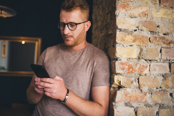 Millennial hipster guy in optical spectacles for provide vision correction using modern smartphone technology for chatting and messaging, young male blogger browsing social networks and web media