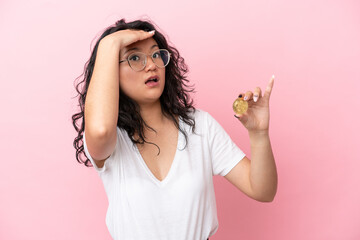 Young asian woman holding a Bitcoin isolated on pink background doing surprise gesture while...