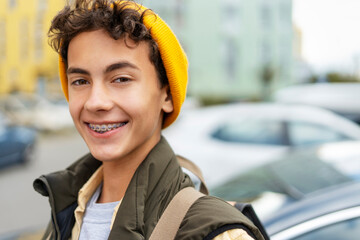 Portrait attractive smiling millennial boy wearing yellow hat, with dental braces on teeth looking at camera on urban street - obrazy, fototapety, plakaty