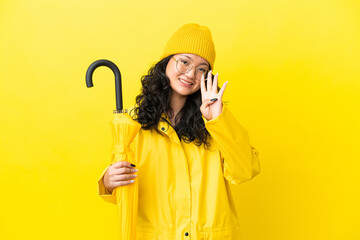 Asian woman with rainproof coat and umbrella isolated on yellow background happy and counting four...