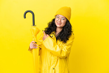 Asian woman with rainproof coat and umbrella isolated on yellow background pointing to the side to...