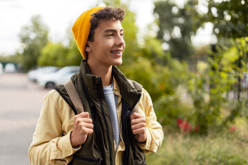 Attractive smiling boy teenager wearing yellow hat with dental braces holding backpack walking to...