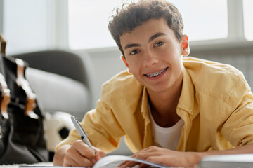 Portrait of attractive smiling boy with dental braces studying, learning language, taking notes lying on floor looking at camera. Education concept - obrazy, fototapety, plakaty