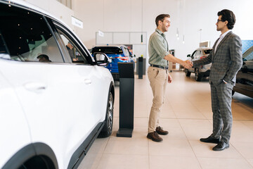 Remote side view of confident car salesman in business suit and cheerful male customer in auto...