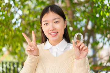 Young Chinese woman holding invisible braces at outdoors smiling and showing victory sign