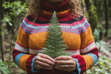 Cut out portrait of unrecognizable woman in nature wearing colorful sweater and holding little tropical leaf against her heart and body. People in love for nature outdoors. Travel healthy lifestyle - obrazy, fototapety, plakaty