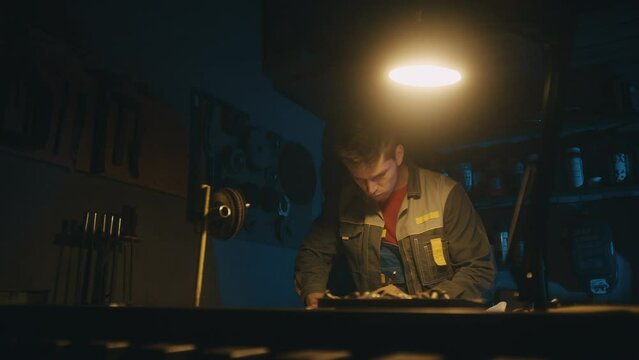 A male engineer works with drawings lying on a steel table in a dark workshop with full equipment, using a pencil. Working indoors on new product prototype in metal manufactory. 4K