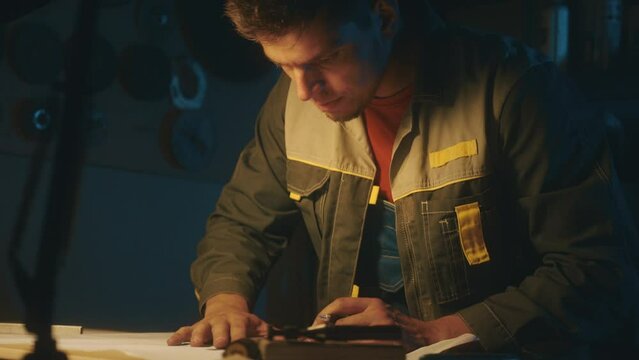 A metallurgical industry worker works on a prototype drawing on a steel table in a dark workshop with full equipment. Engineer working indoors on new product prototype in metal manufactory. 4K