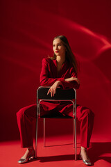 Fashionable confident woman wearing trendy red suit blazer, classic trousers, metallic silver color shoes, sitting on chair, posing on red background. Full-length studio fashion portrait