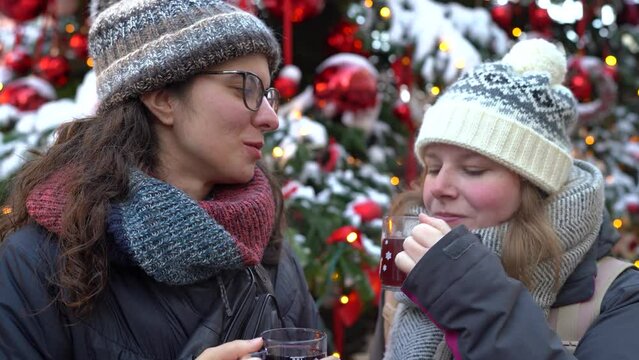 Two girls sharing wine at the christmas market and talking with a big christmas tree behind them