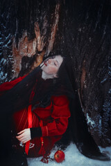 Portrait of a young girl dressed in a red folklore costume, whose face is covered with a black translucent veil. A girl is sitting in a forest near a tree on a winter day. A fairy tale and a legend.