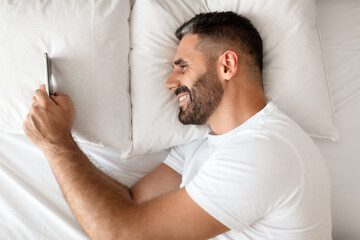 Smiling bearded man using smartphone lying in bed, above view