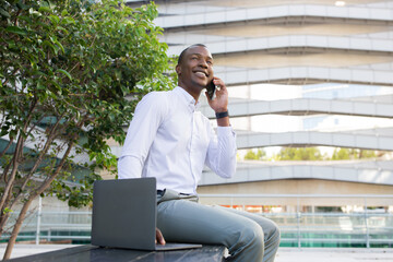Cheerful black businessman in white shirt having pleasant mobile phone conversation outdoors