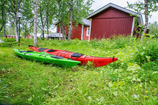 Two kayaks - red and green ones are on the green grass, low level side photo. Small cottage at far background