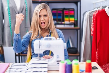 Blonde woman dressmaker designer using sew machine angry and mad raising fist frustrated and...