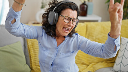 Middle age hispanic woman listening to music sitting on sofa dancing at home