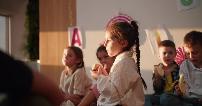 A little girl with a braided hairstyle in a white shirt eats an apple at recess and during lunch in a club for preparing preschool children for school