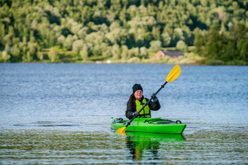Fototapeta na wymiar Mature women in green safety life jacket kayaking in green kayak, she looks at camera. Front side photo on still water with blurry green mountain forest background. Sweden.