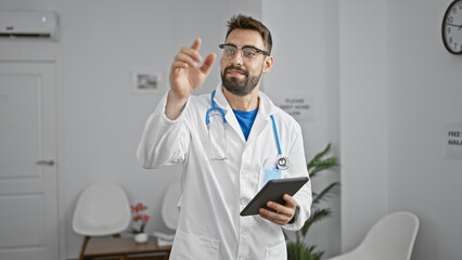 Confident young hispanic male doctor with beard in uniform, smiling and pointing finger at someone in clinic's waiting room, asking for the next turn while professionally using touchpad.