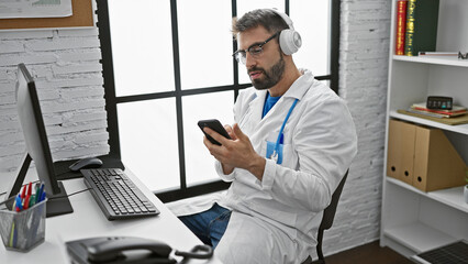 Handsome young hispanic guy, a healthcare professional in his clinic, engrossed in listening to music on headphones while texting on his smartphone.