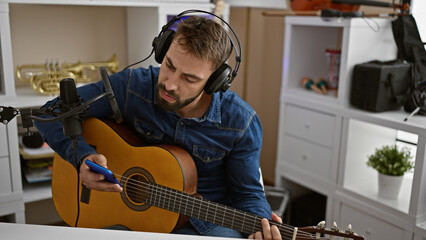 Handsome young hispanic man sitting in a music studio, passionately playing a chord on a classical guitar while looking at his smartphone screen. a melodious artist lost in musical world.