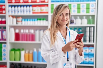 Young blonde woman pharmacist using smartphone working at pharmacy