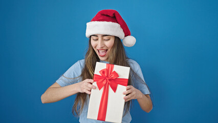 Young beautiful hispanic woman wearing christmas hat holding gift over isolated blue background