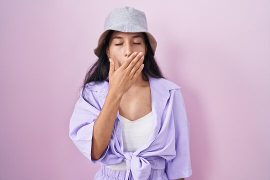 Young hispanic woman standing over pink background wearing hat bored yawning tired covering mouth with hand. restless and sleepiness.