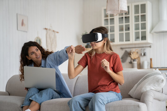 Woman Mother Not Allowing Child To Spend So Much Time In VR, Teen Girl Kid In Vr Headset Sitting On Couch Expanding Virtual World Of Games While Mom Working On Laptop Beside. Virtual Reality And Kids