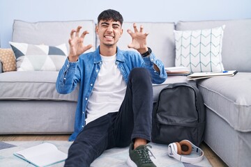 Young hispanic man sitting on the floor studying for university smiling funny doing claw gesture as cat, aggressive and sexy expression