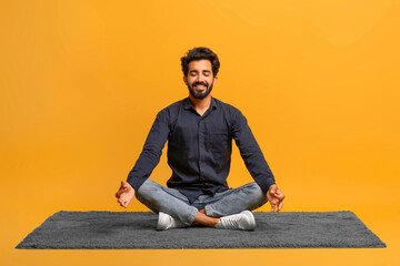 Relaxed young indian man in casual clothes meditating alone, sitting cross-legged on carpe