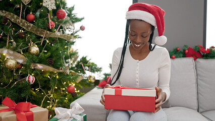 Obraz na płótnie Canvas African american woman holding gift sitting on sofa by christmas tree at home