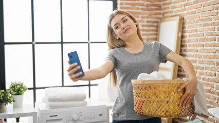 Young blonde woman make selfie by smartphone holding basket with clothes at laundry room