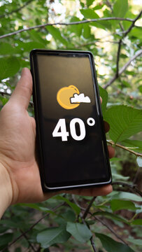 Smartphone thermometer displaying 40 degrees Celsius, indicating warm temperature during a hot summer heatwave weather in Europe, Earth. Blurred green outdoor background.