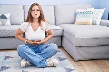 Obraz premium Young caucasian woman sitting on the floor at the living room with hand on stomach because indigestion, painful illness feeling unwell. ache concept.