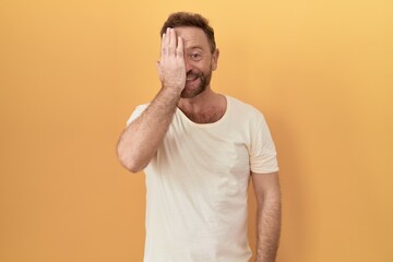 Middle age man with beard standing over yellow background covering one eye with hand, confident smile on face and surprise emotion.