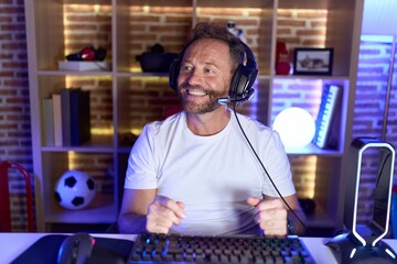Middle age man with beard playing video games wearing headphones looking away to side with smile on...