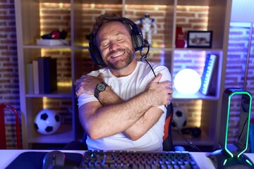 Middle age man with beard playing video games wearing headphones hugging oneself happy and...