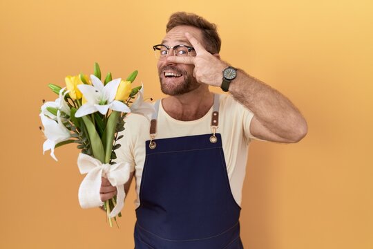 Middle age man with beard florist shop holding flowers doing peace symbol with fingers over face, smiling cheerful showing victory
