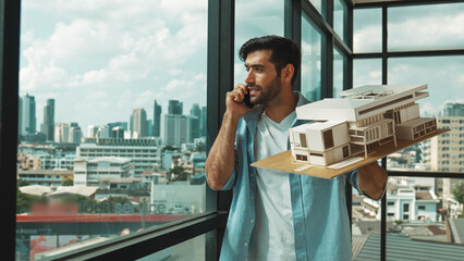 Smart caucasian engineer standing and holding house model while phone calling to manager. Professional manager inspect at architectural model while standing near window. Design concept. Tracery - obrazy, fototapety, plakaty