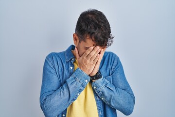 Young hispanic man standing over blue background with sad expression covering face with hands while...