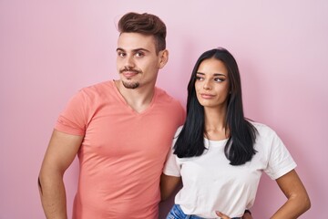 Young hispanic couple standing over pink background smiling looking to the side and staring away thinking.