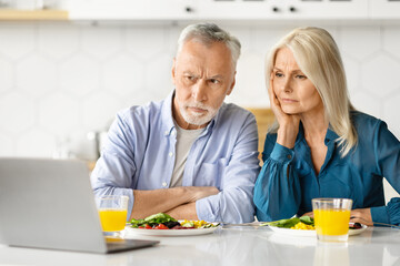 Worried senior couple reading news on laptop during breakfast in kitchen