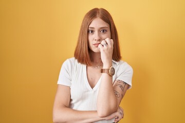 Young redhead woman standing over yellow background looking stressed and nervous with hands on...