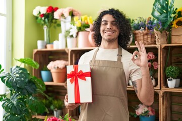 Hispanic man with curly hair working at florist shop holding gift doing ok sign with fingers, smiling friendly gesturing excellent symbol
