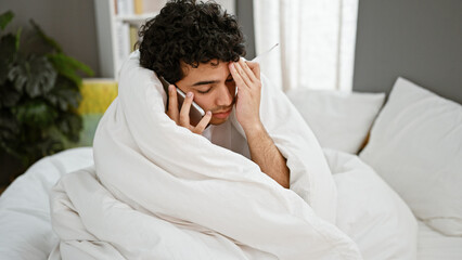Young latin man covering with blanket holding thermometer talking on smartphone at bedroom