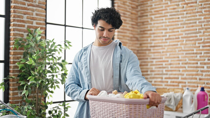 Young latin man smiling confident holding basket with clothes at laundry room