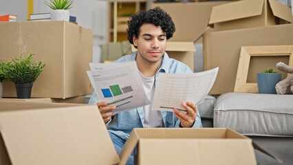 Young latin man reading document sitting on floor looking upset at new home