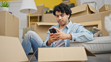 Young latin man using smartphone sitting on floor at new home