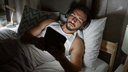 Young arab man using touchpad lying on the bed wearing headphones at bedroom